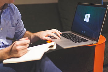 An adult man writing notes while working on a laptop indoors, illustrating a modern work environment. HD wallpaper