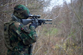 Soldier in camouflage aiming a rifle in a dense forest setting during daylight. HD wallpaper