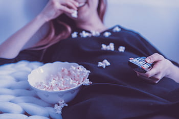 Woman relaxing on couch eating popcorn while watching TV, holding remote control, indoors. HD wallpaper