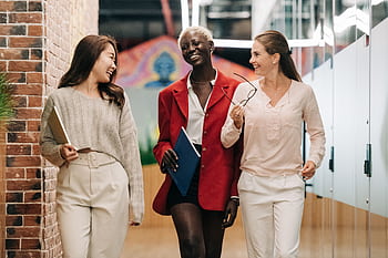 Three diverse businesswomen walking together in a modern office hallway, smiling and talking. HD wallpaper