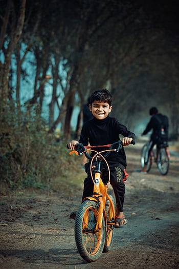 Happy child rides an orange bicycle on a rural dirt path surrounded by trees in Bangladesh. HD phone wallpaper