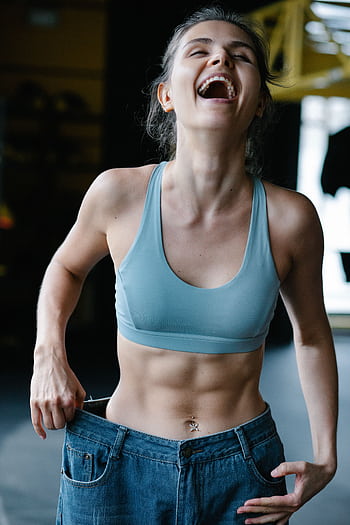 Fit young woman happily shows off weight-loss success by wearing oversized jeans in a gym. HD phone wallpaper
