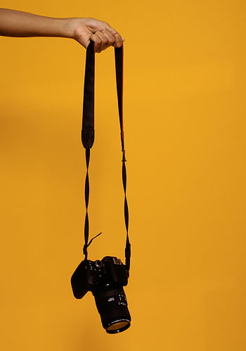 A DSLR camera suspended by hand against a vibrant orange backdrop, showcasing graphy technology. HD phone wallpaper