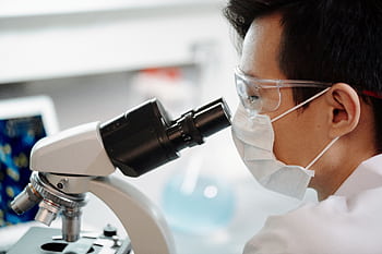 Scientist wearing face mask and goggles examining samples through a microscope in a lab. HD wallpaper