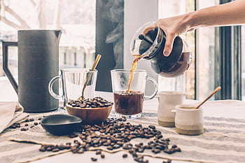 A hand pours freshly brewed coffee into a mug surrounded by coffee beans. HD wallpaper