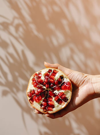 Pomegranate half held against warm shadows, showcasing fresh seeds. HD phone wallpaper