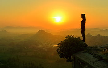 A woman stands silhouetted against a vibrant sunset on a cliff overlooking a misty landscape. HD wallpaper