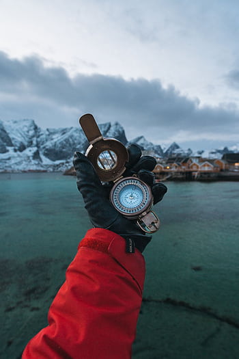 A gloved hand holds a compass in Reine, Lofoten Islands, with a stunning fjord backdrop. HD phone wallpaper