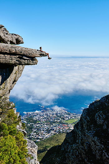 A person sits on a cliff edge enjoying a breathtaking view of the ocean and city below. HD phone wallpaper