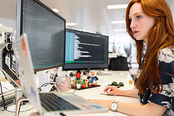 A focused female software engineer coding on dual monitors in a modern office. HD wallpaper