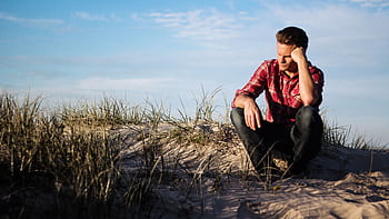 Man in a red plaid shirt sitting thoughtfully on a sandy dune in Australia, surrounded by grass. HD wallpaper