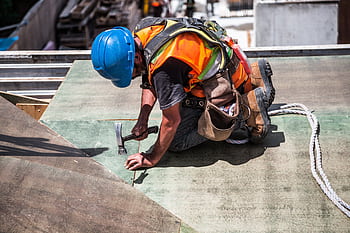 A skilled construction worker in protective gear hammering a rooftop panel. HD wallpaper