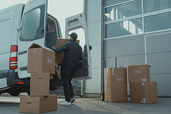 A delivery man unloading cardboard boxes from a van at a warehouse during the day. HD wallpaper