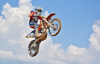 Motocross rider performs an airborne jump under a bright blue sky with clouds. HD wallpaper
