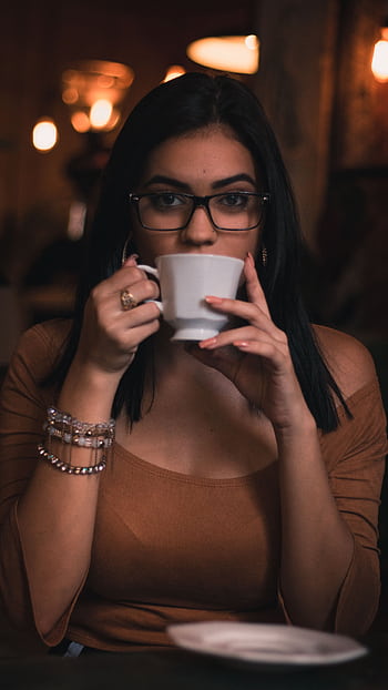 A woman with eyeglasses savoring a cup of coffee indoors at night. HD phone wallpaper
