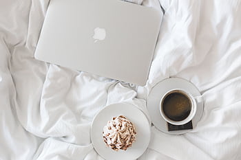 A serene morning setup with a laptop, coffee, and cake on a bed, embodying comfort and relaxation. HD wallpaper