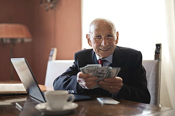 Elderly man smiling while counting cash at a home office setting. HD wallpaper