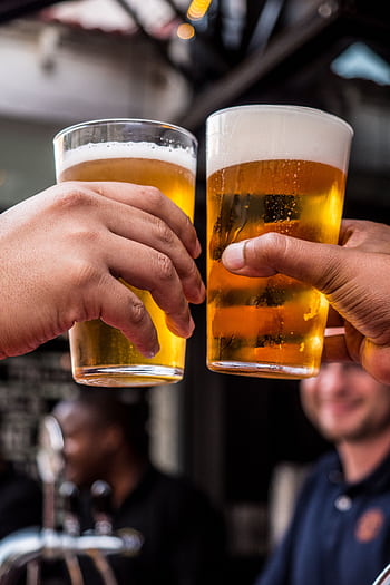 Close-up of two friends toasting with cold beer glasses at a lively bar, celebrating friendship and good times. HD phone wallpaper