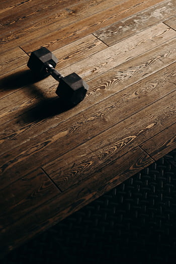 A solitary dumbbell rests on a wooden gym floor, offering ample copy space. HD phone wallpaper
