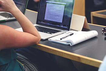 A student in a classroom setting using a laptop and taking notes in a notebook. HD wallpaper
