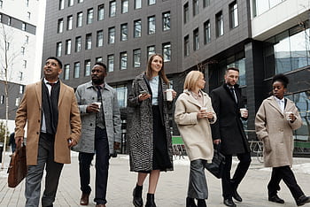 A diverse group of business professionals walking outdoors with coffee cups, showcasing teamwork. HD wallpaper