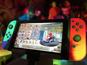 Close-up of a Nintendo Switch showing Mario Kart selection screen, gaming atmosphere in the background. HD wallpaper