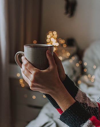Hands holding a warm mug indoors with soft bokeh lights, creating a cozy winter atmosphere. HD phone wallpaper