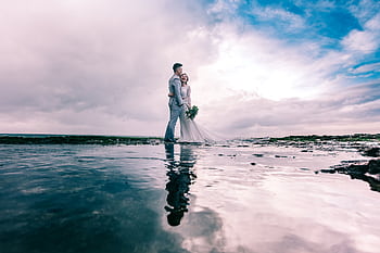 A joyful couple embracing on the beach in wedding attire under a dramatic sky. HD wallpaper