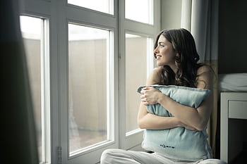 A woman sits by the window, hugging a pillow while smiling warmly. HD wallpaper