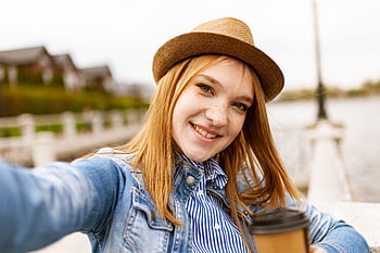 Young woman in a fedora hat takes a selfie with a coffee by the lake. HD wallpaper