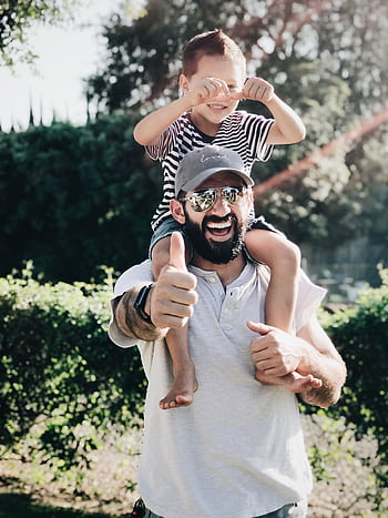Father giving son a ride on shoulders, both laughing and smiling in a park setting. HD phone wallpaper
