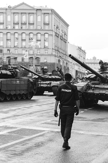 Monochrome of a police officer walking amidst military tanks in an urban setting. HD phone wallpaper