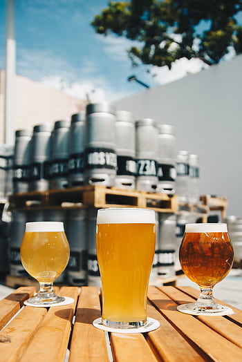 Assorted beer glasses on a wooden table with brewery kegs in the background. HD phone wallpaper
