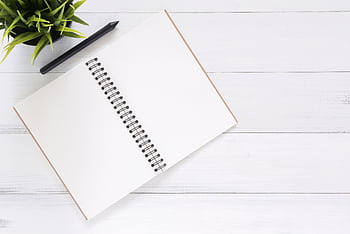Open spiral notebook on white desk with black pen and green plant. Ideal for planning. HD wallpaper