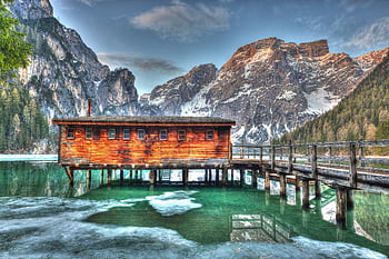 A captivating landscape of a rustic cabin perched over a crystal-clear mountain lake with stunning peaks in the background. HD wallpaper