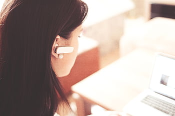 A businesswoman using a Bluetooth headset while working on her laptop in a modern office. HD wallpaper