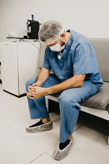 A male doctor in hospital attire sitting pensively, representing healthcare challenges. HD phone wallpaper