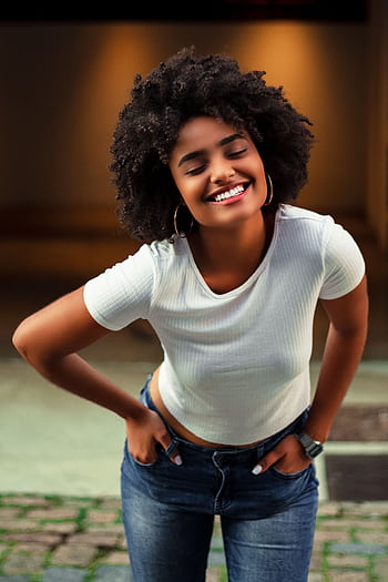 African American woman with afro hair smiling in casual white shirt and jeans, standing outdoors. HD phone wallpaper