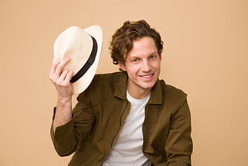 A cheerful man with curly hair tips his hat in a bright studio portrait. HD wallpaper