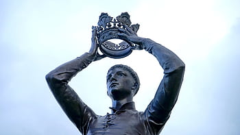 Bronze statue of a figure holding a crown aloft, located outdoors in Warwickshire, England. HD wallpaper