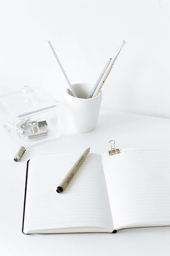 A clean and modern workspace featuring an open notebook, pens, and stationery on a white desk. HD phone wallpaper