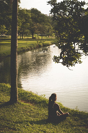 A woman meditates by a serene lake surrounded by lush greenery in a park. HD phone wallpaper