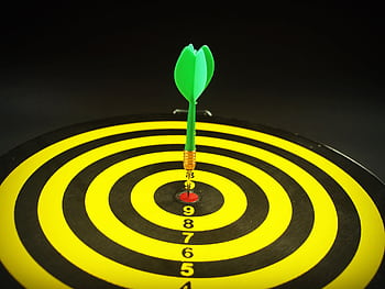Close-up of a green dart hitting the bullseye on a yellow and black target board, symbolizing precision and success. HD wallpaper