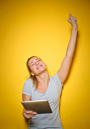 Happy woman with a tablet, celebrating success with arm raised, yellow background. HD phone wallpaper