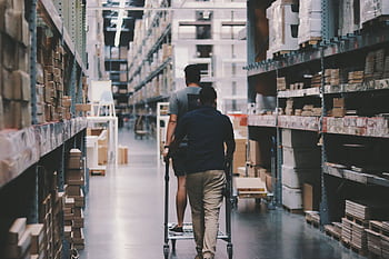 Two men maneuver a trolley in a large warehouse filled with boxes and shelves. HD wallpaper