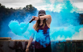 A stylish woman surrounded by bright blue smoke outdoors, creating a dynamic fashion statement. HD wallpaper
