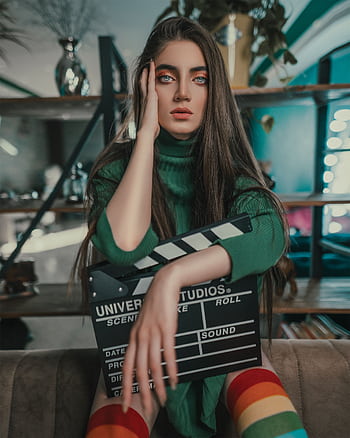 Striking portrait of a model with vibrant makeup and rainbow socks holding a clapperboard indoors. HD phone wallpaper
