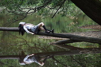 A young woman with a book on her face lies relaxing on a tree trunk by a tranquil lake. HD wallpaper