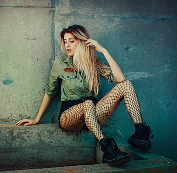 Stylish young woman in fishnet tights sitting against an urban concrete backdrop. HD wallpaper