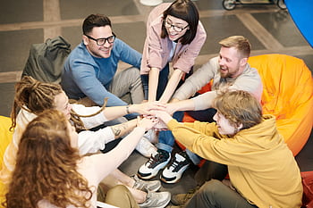 A diverse group of adults sit indoors, joyfully stacking hands together, symbolizing teamwork and camaraderie. HD wallpaper